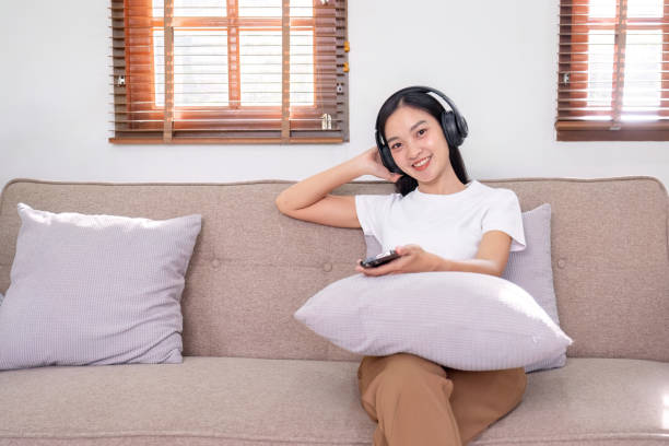 jovem mulher ouvindo música enquanto sentada em um sofá de casa usando fones de ouvido se sente feliz e relaxada passando o tempo livre em casa. - room service audio - fotografias e filmes do acervo