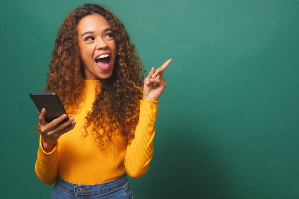 istock Beautiful young woman points thumb to copy space left, cellphone, green studio 1674137227