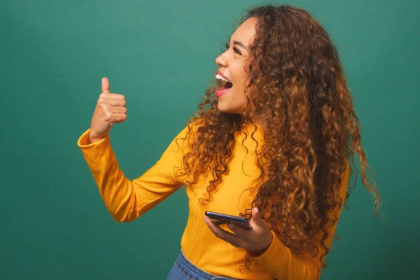 istock Beautiful young woman points thumb to copy space left, cellphone, green studio 1674139156