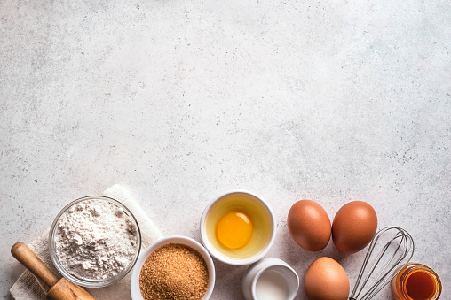 Wheat flour, eggs, rolling pin and whisk on white concrete background, top view, copy space. Ingredients for bakery, baking or cooking concept.