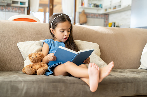 A girl with bare legs and feet reading a fairy tale story book, sitting with her teddy on a couch