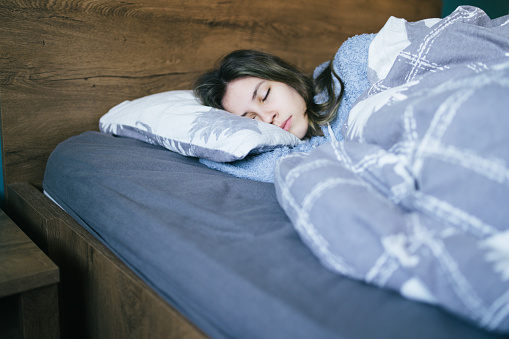 A young woman is sleeping peacefully under a quilt against blue bedding with a wooden headboard