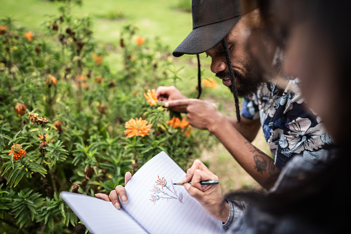 Young man observing flowers while his friend draws them in her notepad on the public park