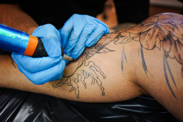 istock Hands of a tattoo artist wearing protective gloves and holding a machine while creating a picture on a man's leg 1674183680