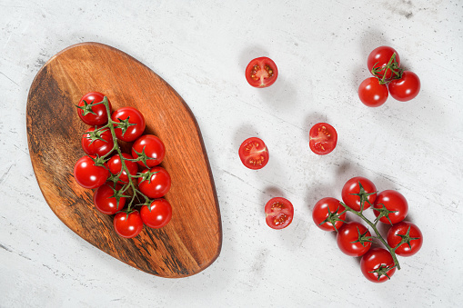 Vibrant small red tomatoes with green vines on wooden chopping board, white stone table under, view from above
