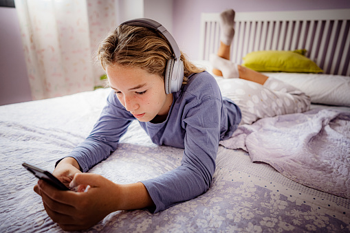 Teenager girl using headphones and text messaging on phone laying face down on her bed. High resolution 42Mp studio digital capture taken with SONY A7rII and Zeiss Batis 25mm F2.0 CF lens