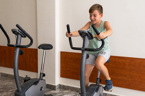 Boy is riding an exercise bike at home gym. Cardio training, leg exercises. Happy family enjoys weekend together in home gym. All family members doing exercise. Healthy lifestyle. Sport and training. Sport and fitness equipment.