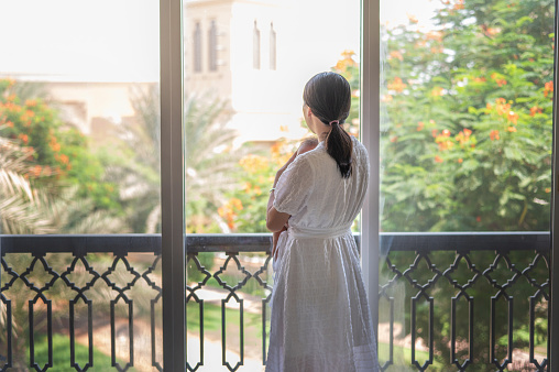 Asian young mother in a white dress looking out the window of her house while holding her newborn son in her arms