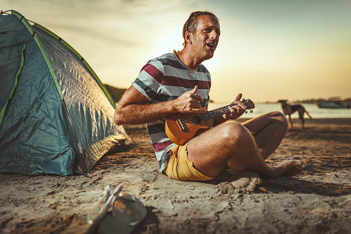 Happy young musician has a great time at the beach. He is sitting by the river, playing ukulele and singing at sunset.