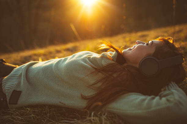 Cute smiling young woman, with headphones on her head, is lying down on the ground in early spring sunny day and listening music.