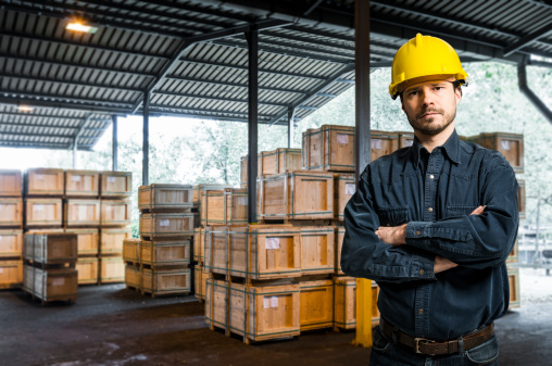 Manager, Inspector, or Mid Adult worker with yellow helmet is standing against covered industrial external warehouse with wooden boxes, packages in medium size.