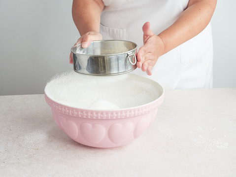 woman's hands in white apron sifting icing sugar with metallic sifter over large pink ceramic bowl on table