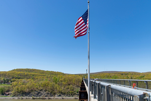 Poughkeepsie, NY, USA-May 2022; Side view of the 1.28 miles Walkway Over the Hudson State Historic Park Bridge from 1889 with American flag in forefront, part of the Hudson Valley Rail Trail