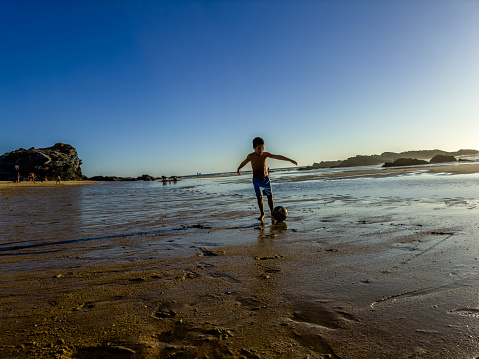 In summer time on a beach at low tide, a boy plays in the sand with a ball in an artistic way