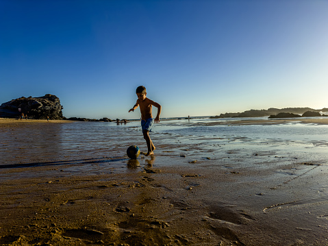 In summer time on a beach at low tide, a boy plays in the sand with a ball in an artistic way