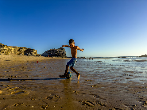 In summer time on a beach at low tide, a boy plays in the sand with a ball in an artistic way