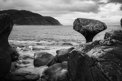 This black-and-white horizontal photograph presents a stunning view of Kannesteinen Beach in Norway, most notably featuring its iconic mushroom-shaped rock. The foreground is covered with sea-eroded stones leading the eye towards the mushroom rock in the middle ground. The calm sea and distant mountain fill the background, all under a dramatically cloudy sky. Captured during a cloudy summer morning, the photograph exudes a sense of stillness and drama.