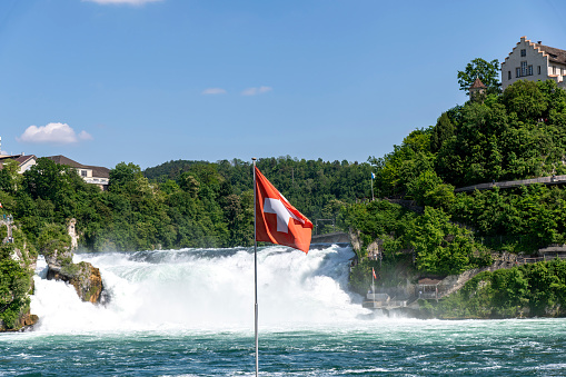Swiss flag with in background a panoramic view over the Rhine falls (Rheinfalle) near Schaffhausen, Switzerland against a white clouded blue sky