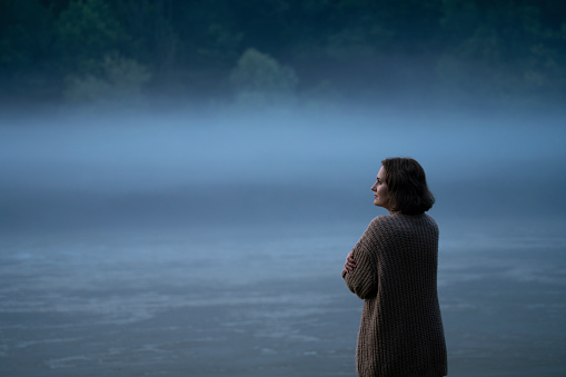 A woman in a sweater looks at the foggy river