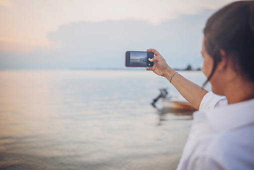 Woman standing on the beach by the sea, she is using smart phone to take pictures of the sea.