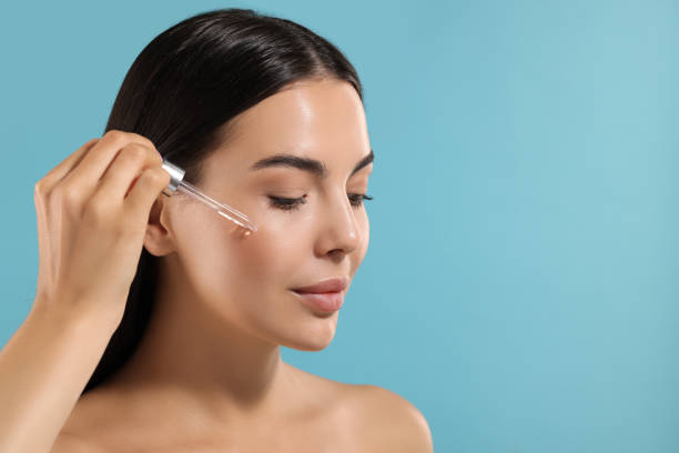 istock Beautiful young woman applying serum onto her face on light blue background. Space for text 1674583363