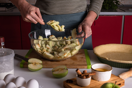Woman mixing cutted apples in a bowl with spices, cinnamon, anice, sugar, cooking apple pie in the kitchen surrounded by other ingredients, Thanksgiving tart preparation, autumn bakery