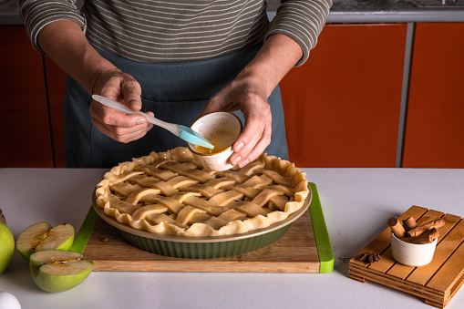 Woman spreading eggs on the raw apple pie with a cooking brush in a bakery dish, ready to be putting in the oven, Thanksgiving tart preparation, autumn bakery. Crispy weather sweets. Recipe