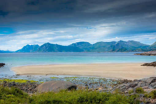 This is an image of a coastal scene where a pebbly beach meets a calm, blue sea. Gentle waves are lapping onto the shore. In the foreground, there is some greenery and what appears to be a disused structure, possibly a broken wooden fixture or bench. In the distance, the coastline extends into a series of headlands or foothills, each progressively fading into a blue haze. The sky is clear with only a few wispy clouds, and the overall impression is one of a tranquil, sunny day by the sea.