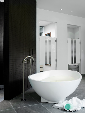 Bathtub freestanding by wall of mosaics. Water tap comes up from gray tiled  floor. In background toilet room and wardrobe, both hidden behind bar-like swinging doors. Bathtub in white corian.