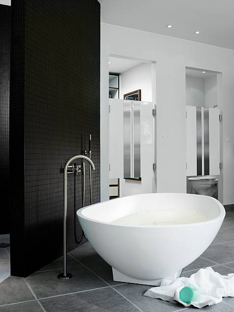 Luxury bathroom Bathtub freestanding by wall of mosaics. Water tap comes up from gray tiled  floor. In background toilet room and wardrobe, both hidden behind bar-like swinging doors. Bathtub in white corian. drenched stock pictures, royalty-free photos & images