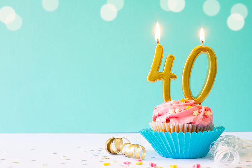 Vanilla buttercream cupcake with pink icing, sprinkles and gold 40th birthday candles. \nShot  on a light blue background with defocused lights.