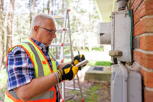 Mature gray haired engineer worker in glasses and vest, writing notes to document his findings of gas meters and pipes after storm outdoors