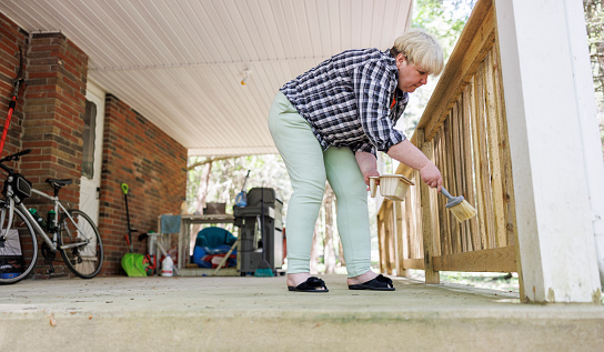 Mature woman in plaid shirt painting fence, prevent fungus and mold on porch. Surface level view