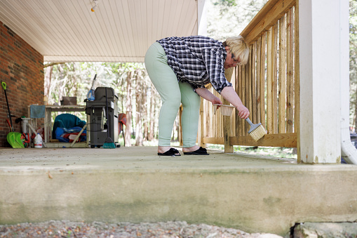 Mature woman bending over, waterproofing wooden fence to maintain its appearance and prevent mold outdoors. Surface level view