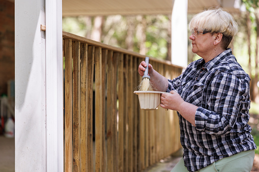 Mature woman with gray hair in plaid shirt waterproofs wooden fence to fend off mold and decay