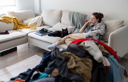 Exhausted young brunette Caucasian woman in eyeglasses resting on couch with black cat in dirty messy living room