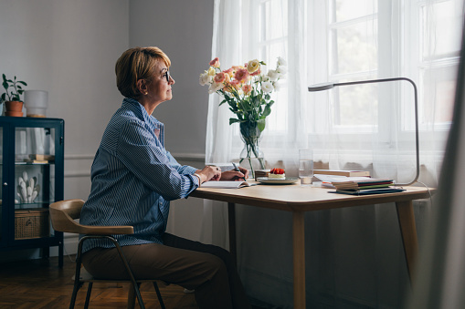 A side view of a pensive focused Caucasian female sitting at the desk and looking out the window while contemplating some ideas to write down in her notebook.