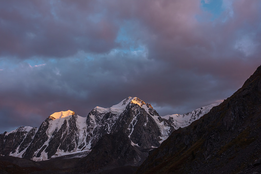 Atmospheric landscape with sunset gold reflection on huge snowy mountain top in violet dramatic sky. Scenic view to giant snow mountains in dusk. Snow-covered mountain range silhouettes in twilight.