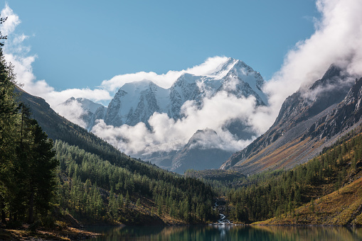 Scenic landscape with turquoise mountain lake in autumn valley against large snow mountains in low clouds in morning sunlight. Alpine lake with view to sunlit high snowy mountain range in low clouds.