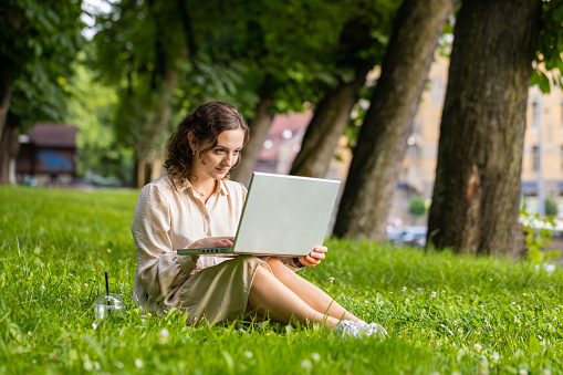 Smiling young woman using laptop typing text answering messages chatting online looking computer screen social media app working. Girl sitting on grass in urban sunset city park holding phone in hands