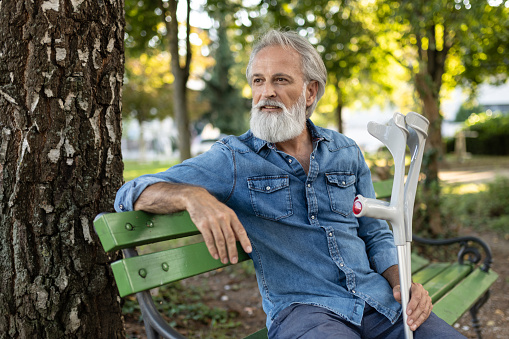 An injured elderly gray-haired man with a beard and crutches sits in the park on a green bench, looks around, greets passers-by with a hand and a phone in his hand. He's talking on his cell phone.