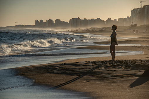 Mature woman standing in front of the sea looks at the waves that break, in the background the mist and buildings.