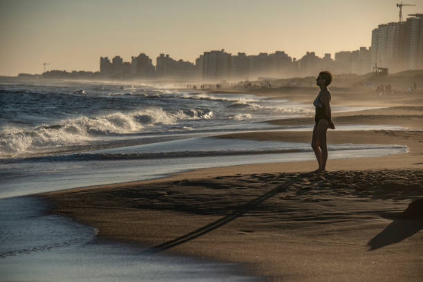 Mature woman standing in front of the sea looks at the waves that break, in Punta del Este, Uruguay. Mature woman standing in front of the sea looks at the waves that break, in the background the mist and buildings. playas del este stock pictures, royalty-free photos & images