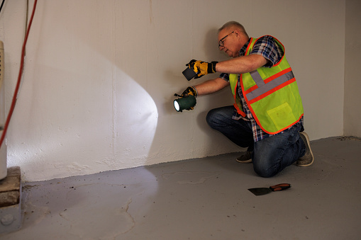 Mature claim agent in glasses using flashlight to examine basement wall after flood. Traces of spillage on the floor due to water damage