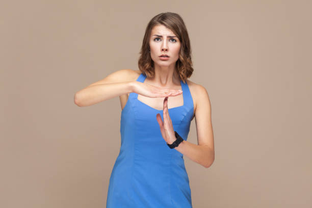 Bossy woman with wavy hair making time out gesture, has serious frustrated expression, needs to stop Portrait of bossy woman with wavy hair making time out gesture, has serious frustrated expression, needs to stop, wearing blue dress. Indoor studio shot isolated on light brown background. time out signal stock pictures, royalty-free photos & images