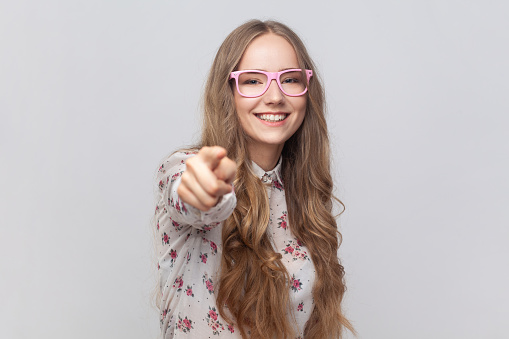 Portrait of pretty cheerful young adult woman in glasses with wavy blond hair looking at camera with toothy smile, pointing finger, suspecting you. Indoor studio shot isolated on gray background.