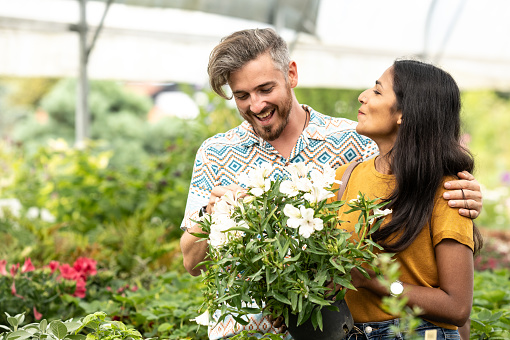 In the greenhouse, a young couple, an Indian woman and a white man, happy and smiling, are buying new plants, new flowers, potted plants for their apartment.