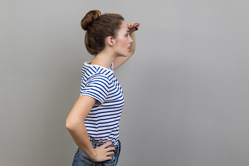 Side view portrait of beautiful ambitious woman wearing striped T-shirt watching far away with hand above eyes, looking forward to future. Indoor studio shot isolated on gray background.