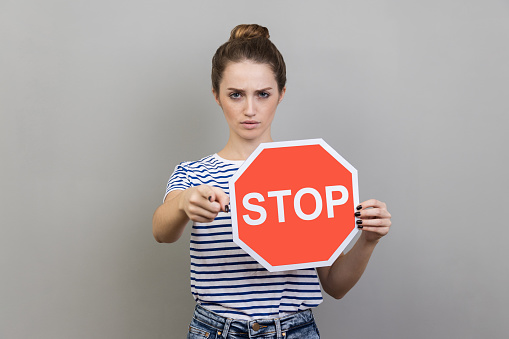 Portrait of serious bossy woman with bun hairstyle wearing striped T-shirt holding red stop sign pointing at camera, has strict expression. Indoor studio shot isolated on gray background.