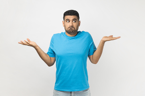 Portrait of puzzled uncertain unshaven man wearing blue T- shirt standing shrugging shoulders, doesn't know how to make decision. Indoor studio shot isolated on gray background.